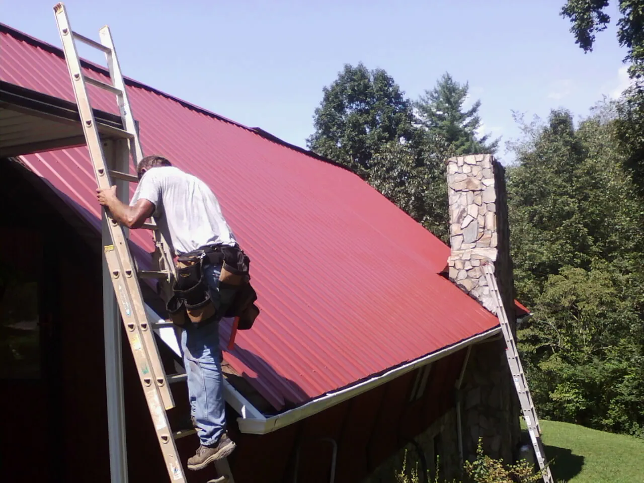 Roofer on a ladder beside a red metal roof and stone chimney — Soffit & Fascia Repair by Roofing Brothers in Sebastian